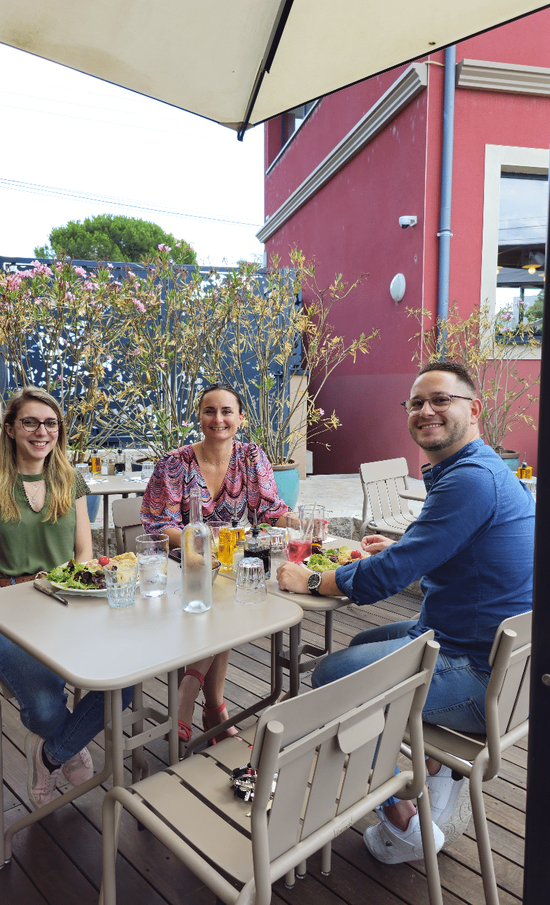 Trois amis souriants attablés en terrasse chez Fabioli, salades et boissons fraîches au soleil