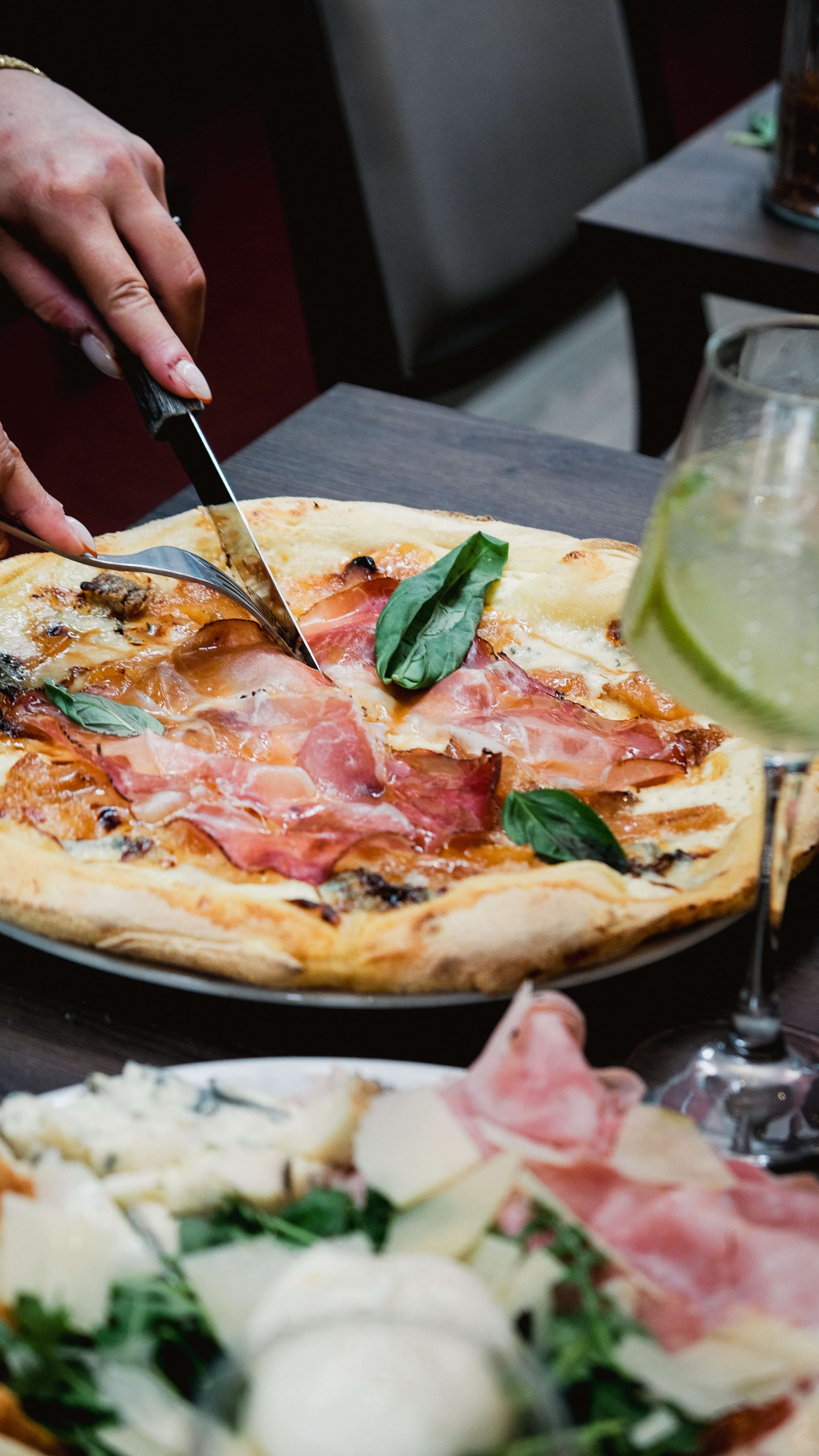 Pizza au jambon cru et basilic découpée à table, avec un spritz et une planche d'antipasti