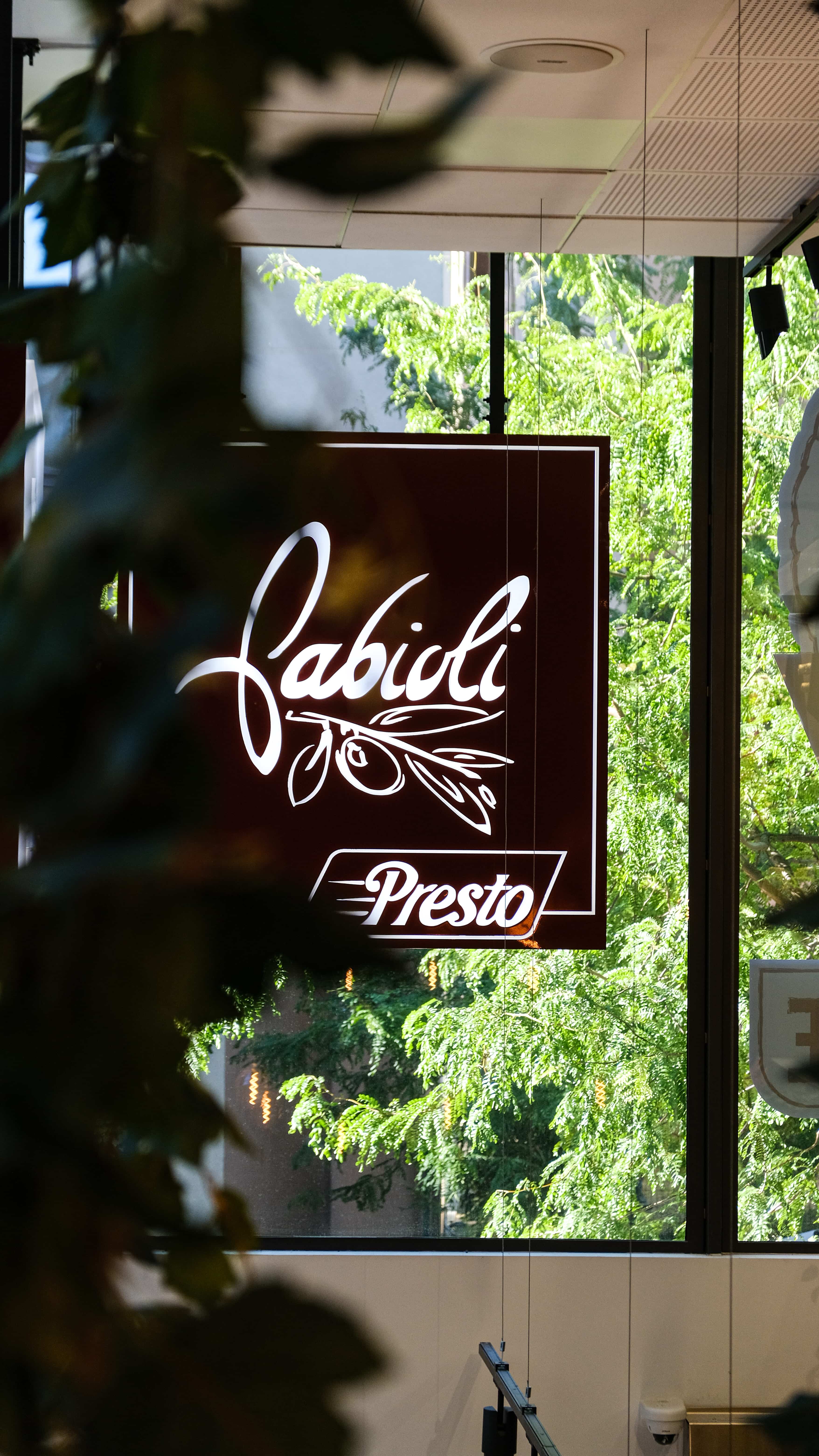 Enseigne Fabioli Presto vue à travers la vitrine, entourée de verdure