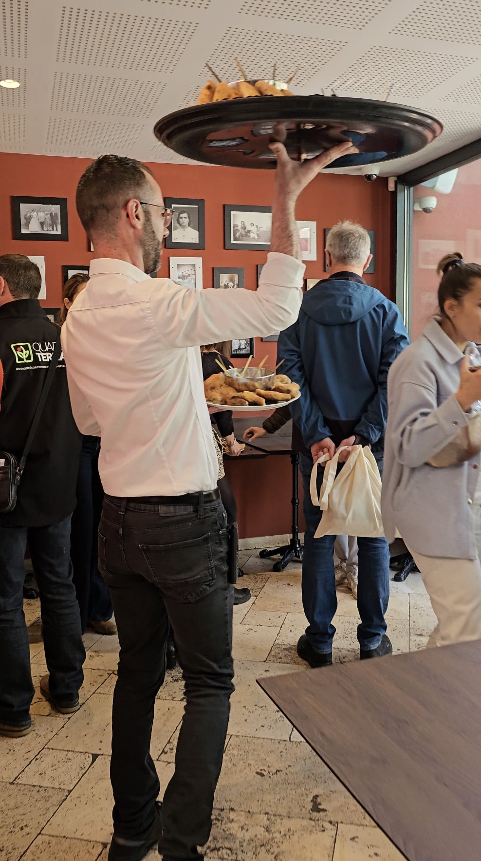 Serveur Fabioli portant un grand plateau de bouchées apéritives au-dessus des convives, devant le mur de photos de famille