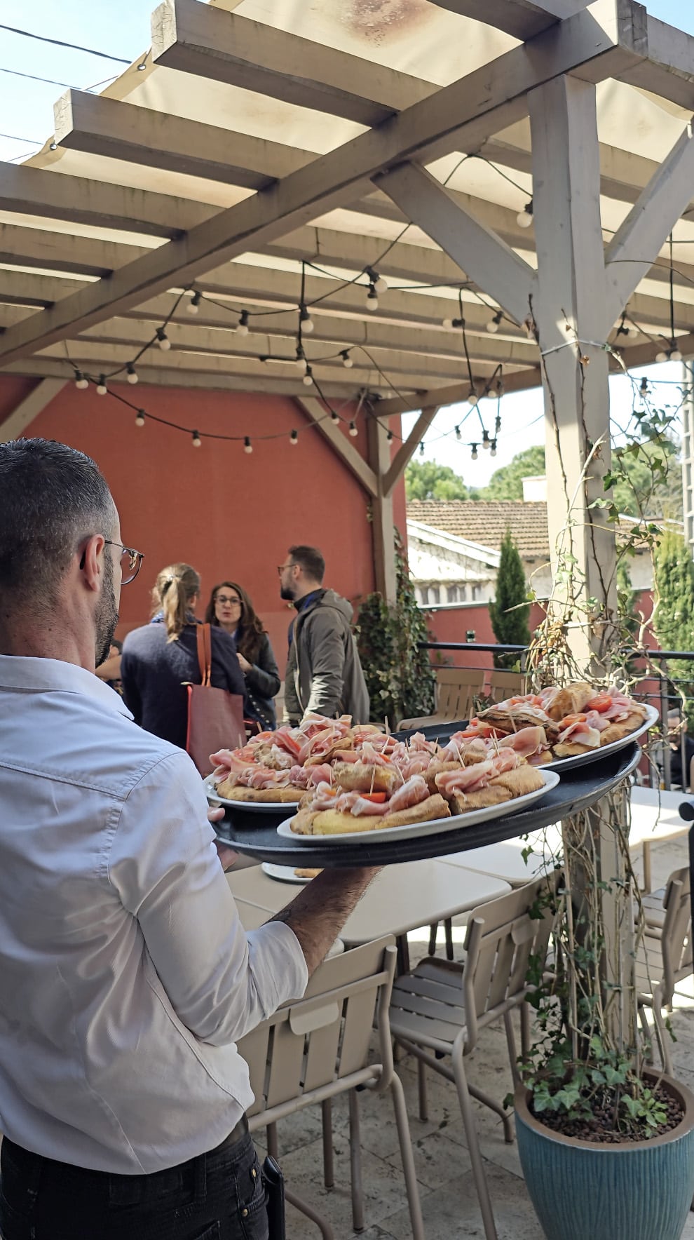 Serveur Fabioli apportant des plateaux de focaccia au jambon cru en terrasse, sous la pergola aux guirlandes lumineuses