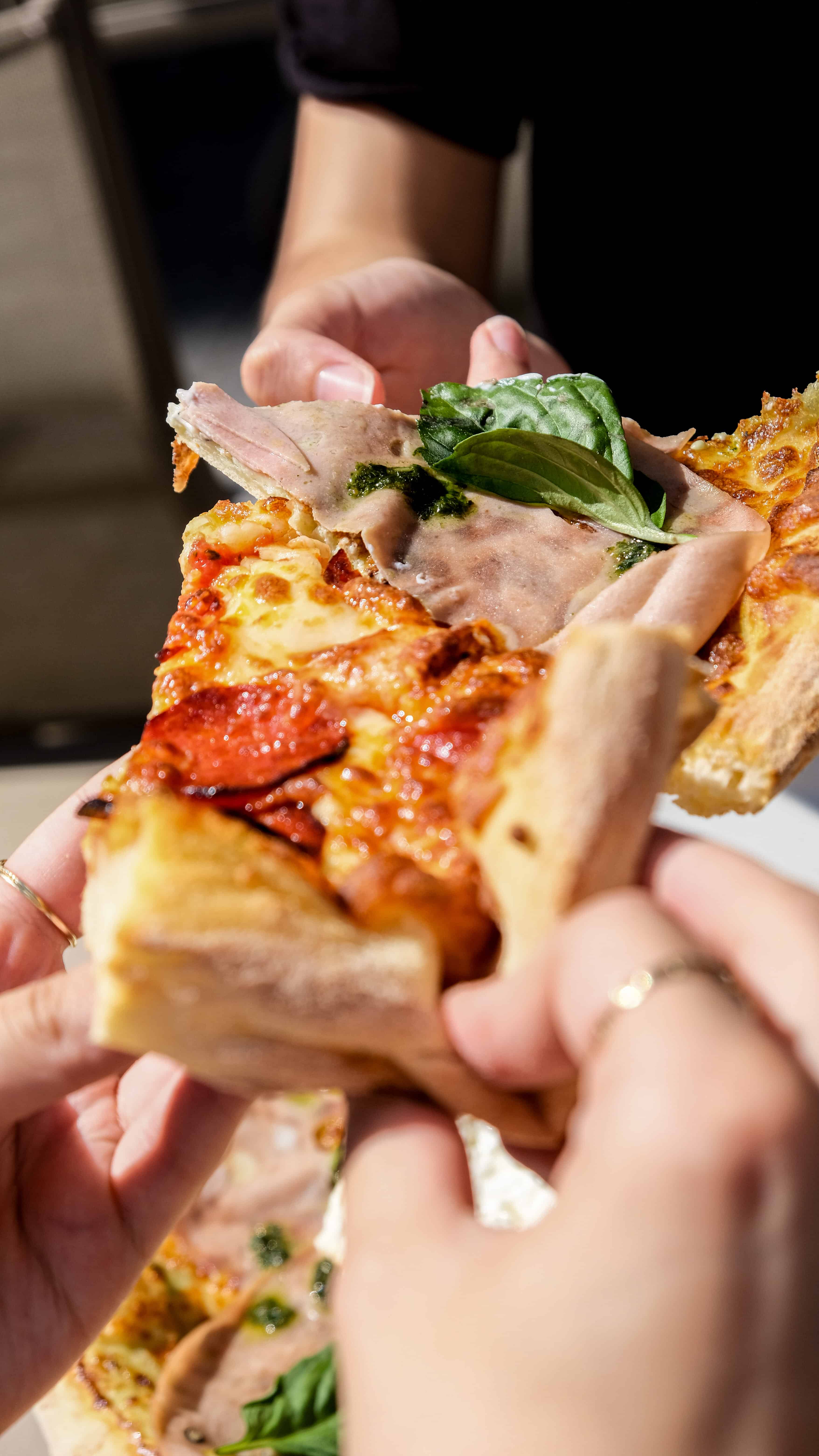 Plusieurs mains se partageant des parts de pizza Fabioli garnies de jambon, pepperoni et basilic
