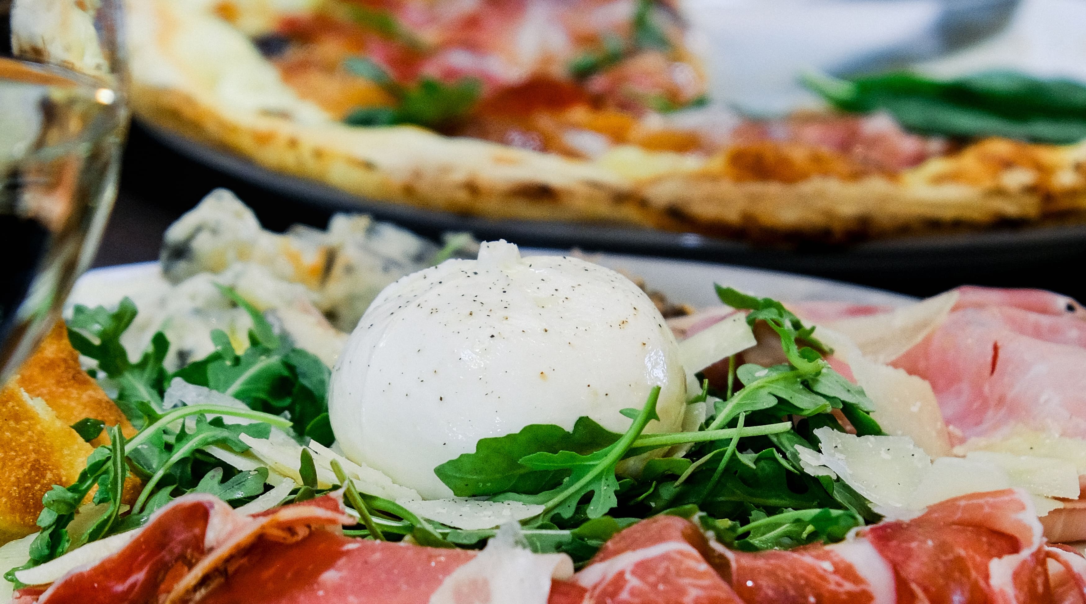 Burrata crémeuse sur un lit de roquette, jambon cru et copeaux de parmesan, pizza en arrière-plan chez Fabioli